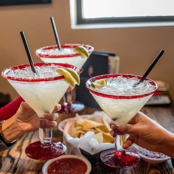 Three hands clinking Picoso's margaritas with red-rimmed glasses, limes, and straws, alongside chips and salsa.
