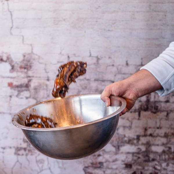 A chef's hand tossing saucy ribs in a metal bowl against a brick wall.