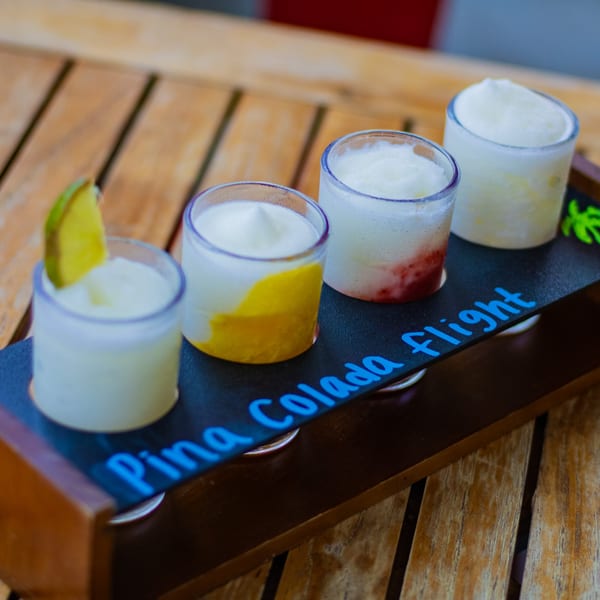 Four Pina Colada flight drinks in small glasses on a wooden tray with a lime wedge.