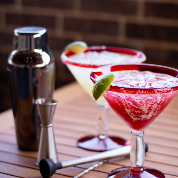 Two Picoso's Mexican Kitchen margaritas, one red and one yellow, with limes and cocktail tools.