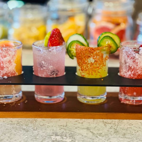 A flight of colorful Picoso's Mexican Kitchen margaritas, garnished with fruit and chili, on a wooden tray.
