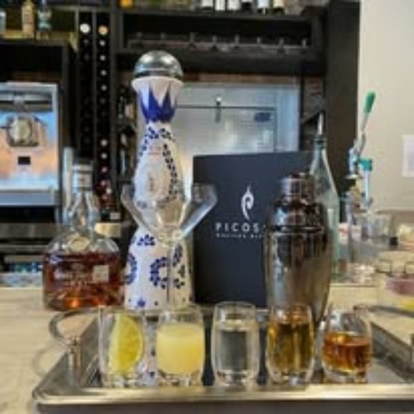 A tequila tasting flight with a decorative bottle, shaker, and Picoso's Mexican Kitchen menu on a bar.